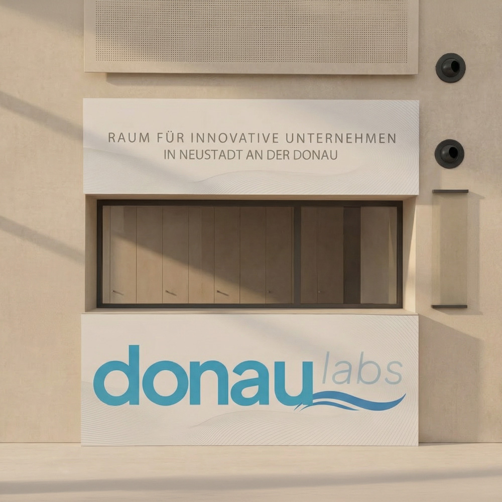 Zwei große,weiße Schilder für einen Empfangstresen für die Firma "Donaulabs". Ein Schild ist in weiß, mit dezenten blau-grauen Wellenlinien und der Aufschrift " RAUM FÜR INNOVATIVE UNTERNEHMEN IN NEUSTADT AN DER DOANU" hängt über einer Öffnung and er Wand. Darunter ist ein weißes Schild befestigt, mit dem großen Logo-Schriftzug "donaulabs".
